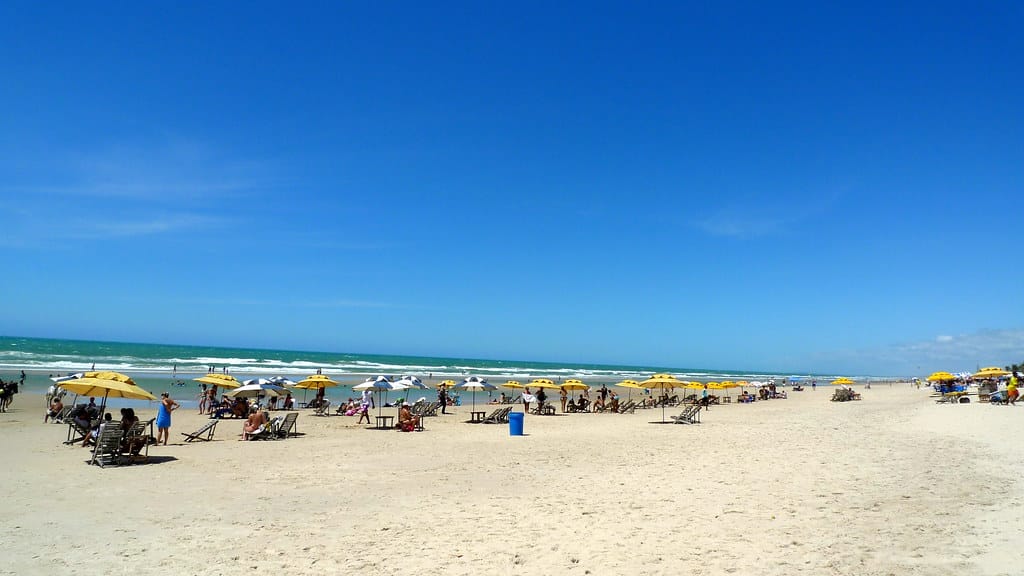 Faixa de areia da praia do Futuro em Fortaleza. Ela está cheia de pessoas com cadeiras de praia e guarda-sóis. Ao fundo é possível ver um mar. 