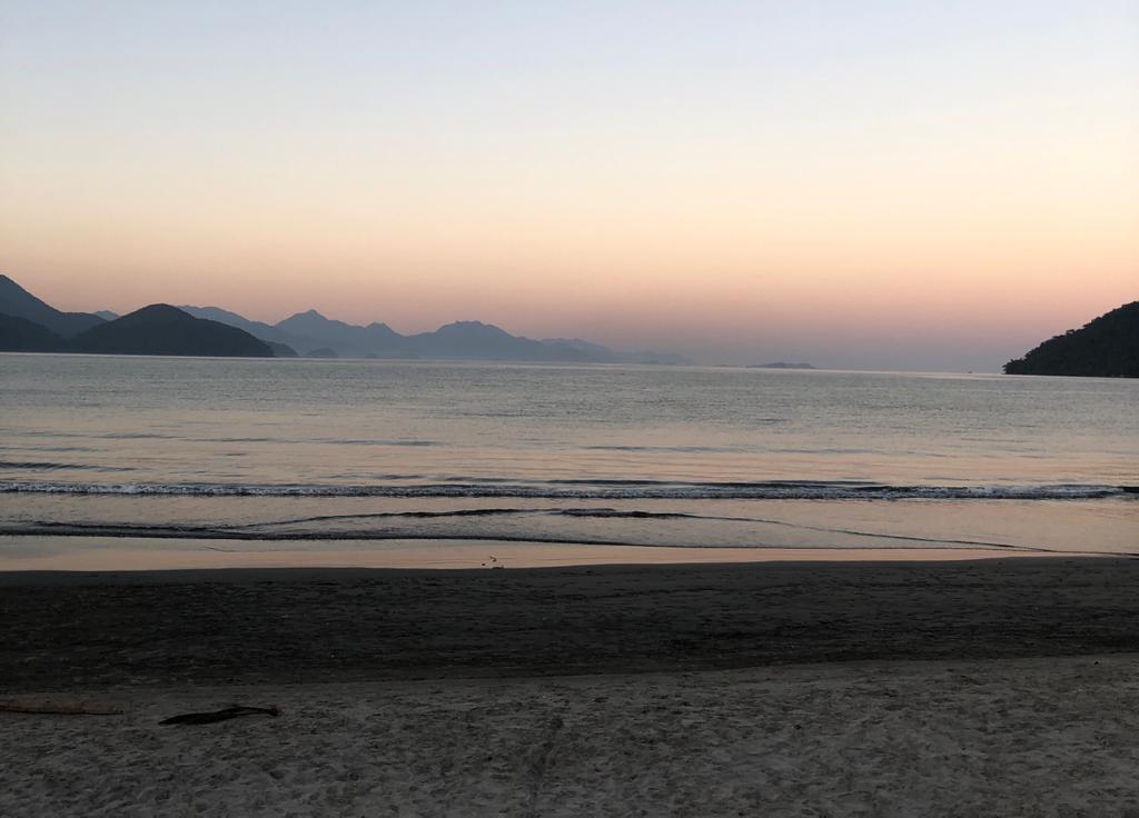 Foto da Praia do Itaguá. Areia na frente e um mar com ondas baixas. No fundo montanhas e um céu rosado com o pôr do sol.
