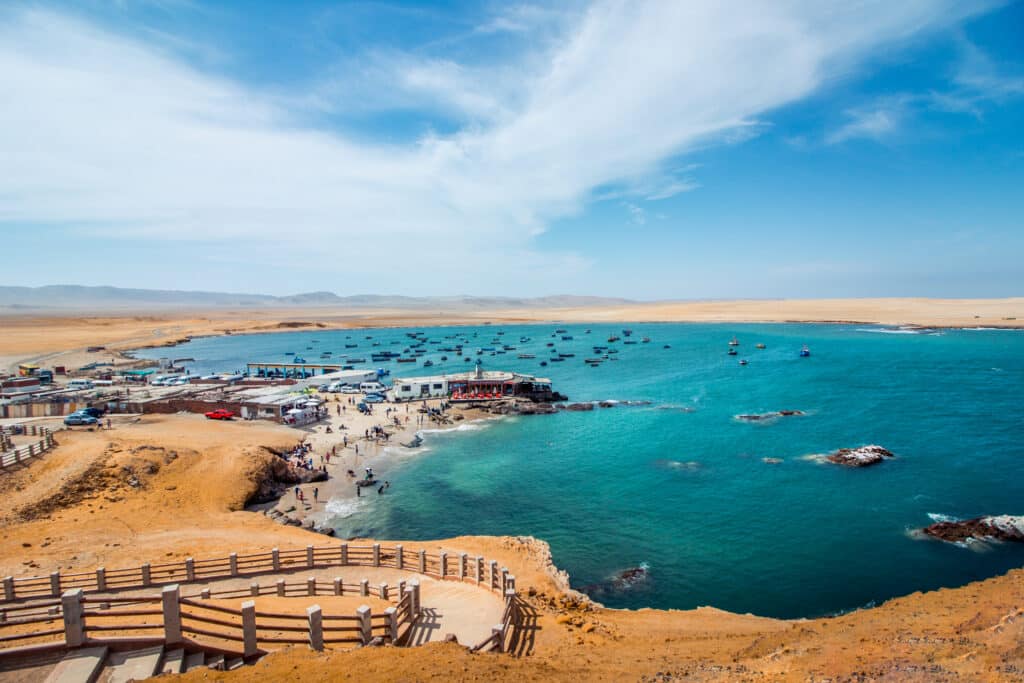 Vista de um mirante do mar de Paracas cercado de falésias amarelas, com barcos navegando e pessoas na orla. Há uma escadaria de acesso, além de carros e casas em uma parte da costa. Representa o que fazer no Peru