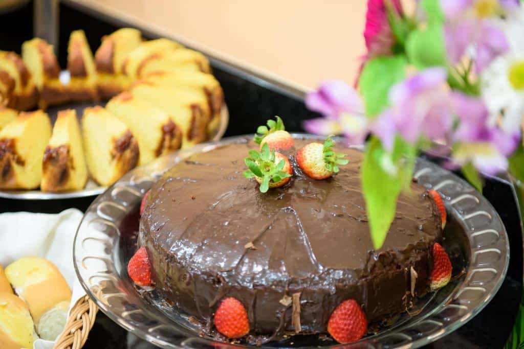 Foto de um bolo de chocolate com morangos no Prisma Plaza Hotel. Atrás, vemos outras fatias de bolos e pães.