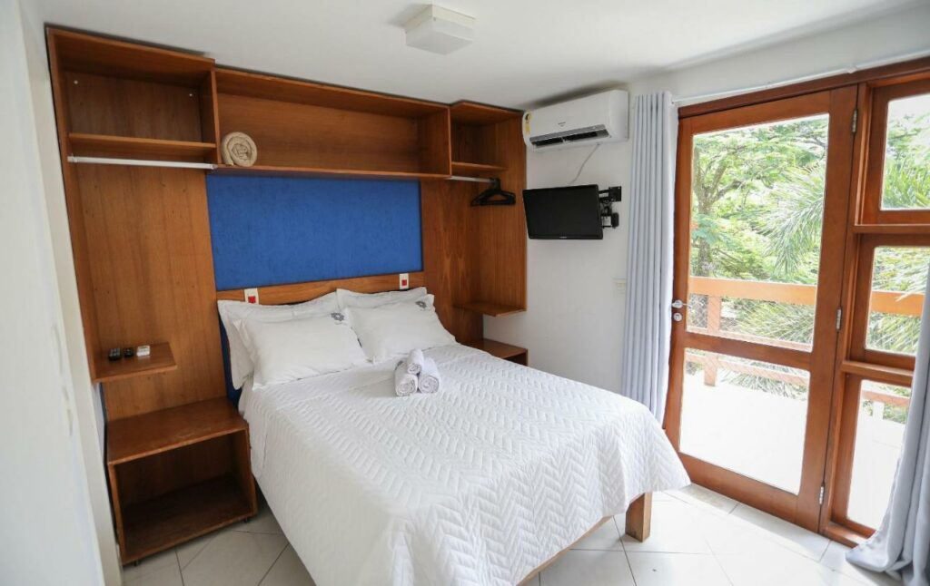 Quarto do Alapage Toque Toque com cama de casal do lado esquerdo da imagem e do lado esquerdo da cama com TV presa na parede. Representa airbnb na praia de Calhetas.