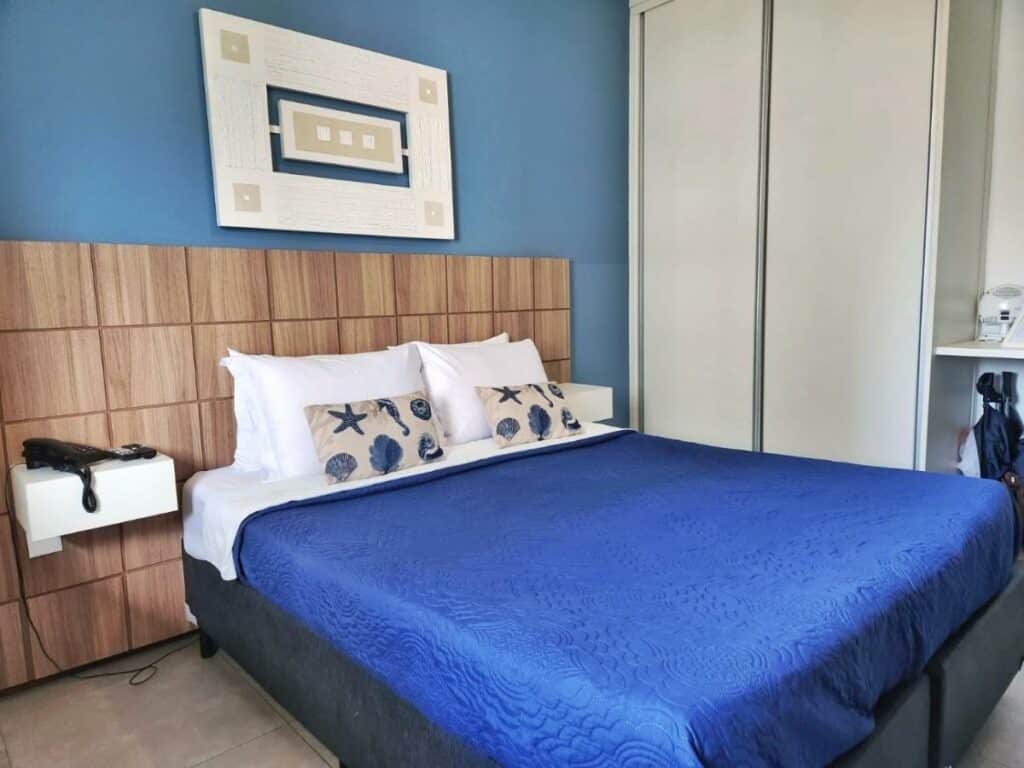 Quarto do Apartamento Barra Pé na Areia com cama de casal do lado esquerdo da imagem com um telefone do lado esquerdo da cama em cima de uma cômoda. Representa airbnb na Barra da Tijuca.