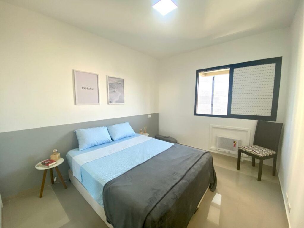 Quarto do Apartamento renovado Barra Bella com cama do lado esquerdo da imagem ao centro do quarto em cada lado da cama uma cômoda em cada lado da cama e do lado esquerdo da cama uma cadeira. Representa airbnb na Barra da Tijuca.