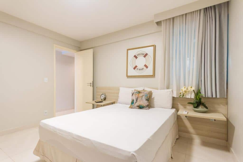 A cama está centralizada no meio do quarto, há uma prateleira em cada lado da cama e no lado direito há uma janela com cortina. No lado esquerdo está a porta que dá acesso ao restante do apartamento. Imagem para ilustrar o post airbnb em Angra dos Reis.