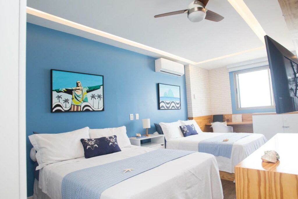 Quarto do Barra da Tijuca de frente para o mar – 2 suítes até 6 pessoas em camas com uma cama de casal do lado esquerdo com uma cômoda do lado esquerdo da cama e do lado esquerdo da cômoda outra cama de casal, e em frente as camas uma cômoda com TV presa na parede.