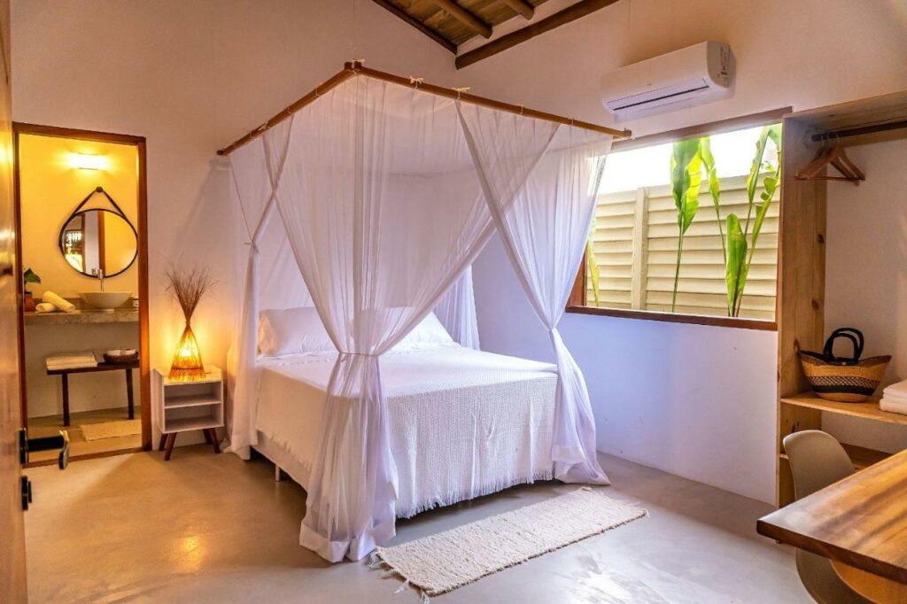 Quarto da Casa Jenipapo Trancoso – Sinta o pedacinho do céu com cama de casal do lado esquerdo da imagem no centro com uma cômoda do lado direito da cama com luminária. Representa airbnb em Trancoso.