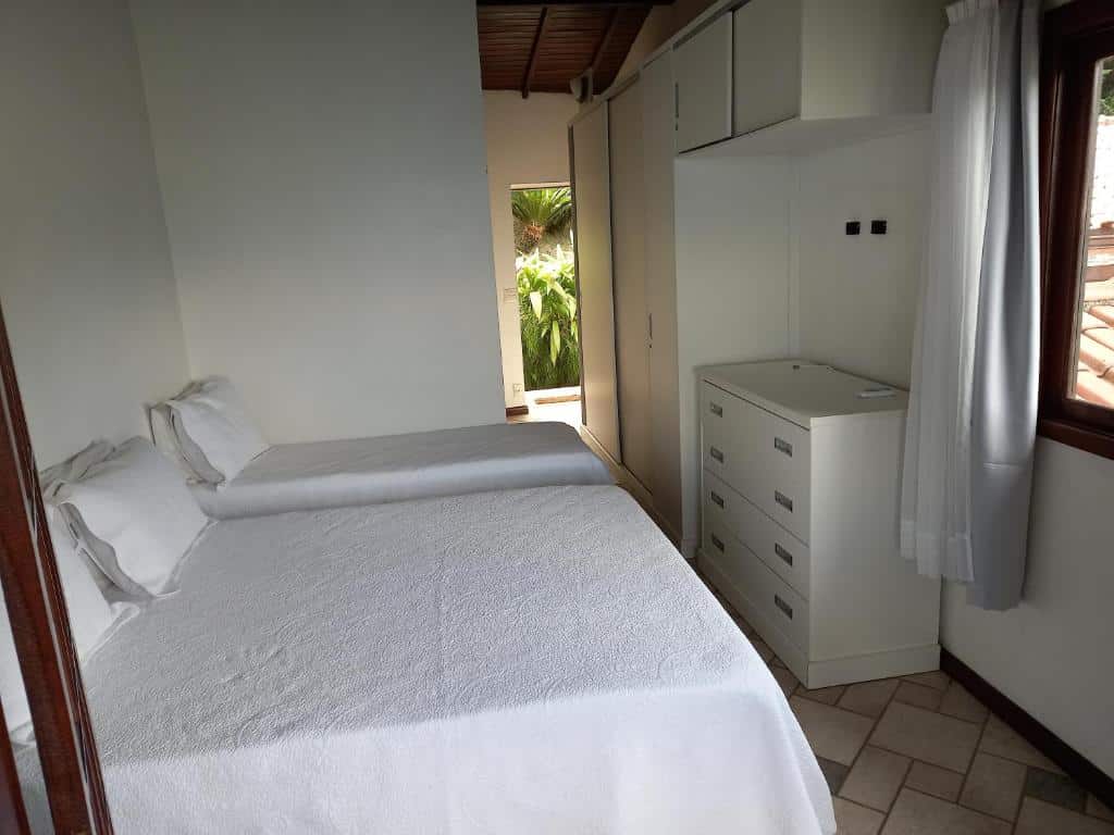 Há duas camas encostadas na parede do lado esquerdo do quarto, uma cômoda e um armário no lado direito, é possível ver um pedaço de uma janela de vidro com cortina e no fundo do quarto há uma porta com vista para um jardim verde. Imagem para ilustrar o post airbnb em Angra dos Reis.