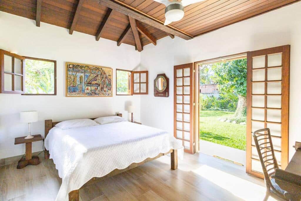 Quarto da Casa na Barra do Sahy com cama de casal do lado esquerdo da imagem em cada lado da cama uma cômoda com luminária em frente a cama uma mesa de trabalho com cadeira. Representa airbnb em São Sebastião.