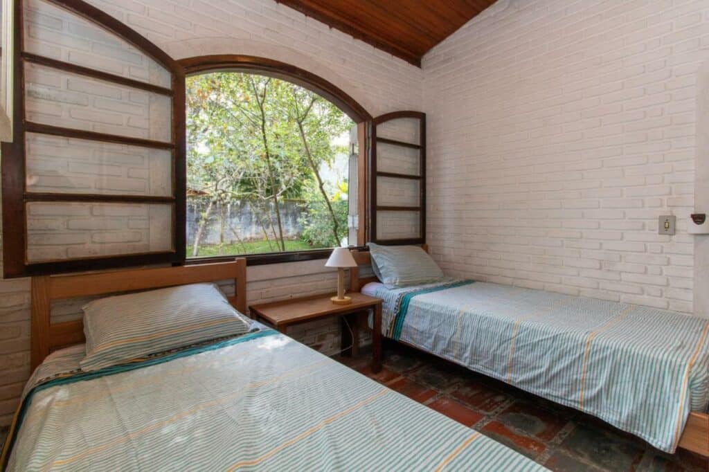 Quarto da Casa Tranquila com duas cama de solteiro em cada lado da cama e ao meio delas uma cômoda com luminária em baixo de uma janela ampla. 
