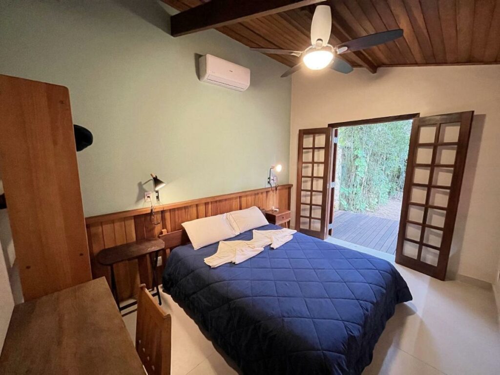 Quarto da Casa Tubarão – A Melhor  com mesa de trabalho com cadeira do lado esquerdo da imagem a frente, já no centro do quarto a cama de casal com uma cômoda em cada lado da cama. Representa airbnb na Barra do Sahy.