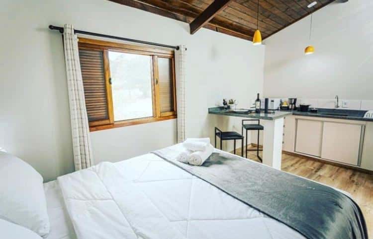 Espaço interno do Chalé Mantiqueira que mostra uma cama de casal, ao lado uma janela de madeira aberta com uma cortina e ao fundo a cozinha compacta.