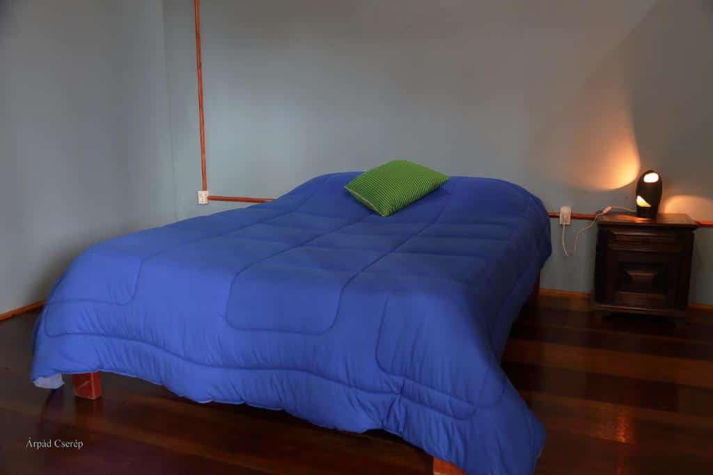Quarto do Chalés Samadhi com uma cama de casal com um cobertor azul e uma almofada em cima. O chão é de madeira e do lado da cama tem uma mesinha com um abajur.