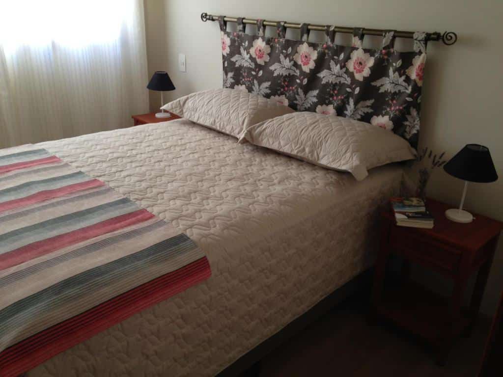 Quarto do airbnb Charme no Centro com uma cama de casal com dois travesseiros e uma colcha. De cada lado da cama tem uma uma mesinha de madeira com um abajur e do lado esquerdo ao fundo tem uma cortina branca.