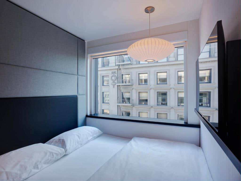 Quarto do citizenM San Francisco Union Square com cama de casal do lado esquerdo da imagem do lado esquerdo da cama janela ampla com vista para a cidade. Representa hotéis em San Francisco.