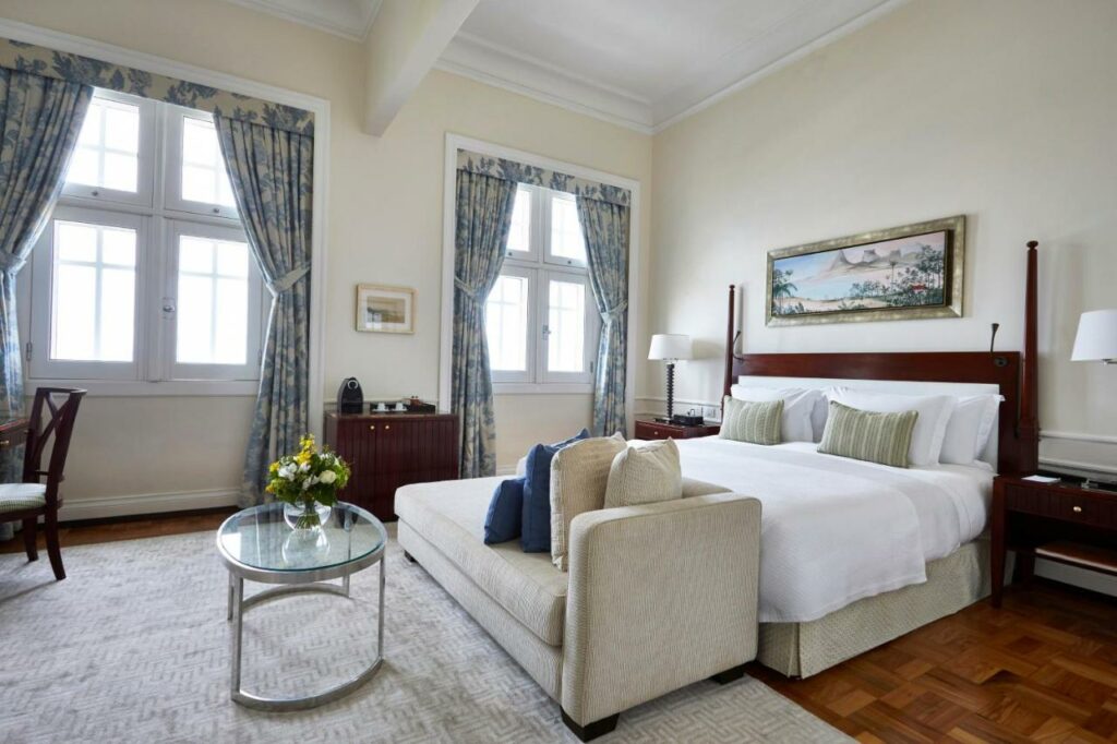 Quarto do Copacabana Palace com cama de casal do lado direito da imagem com uma cômoda em cada lado da cama com luminária, no pé da cama um sofá e em frente ao sofá uma mesa de centro.