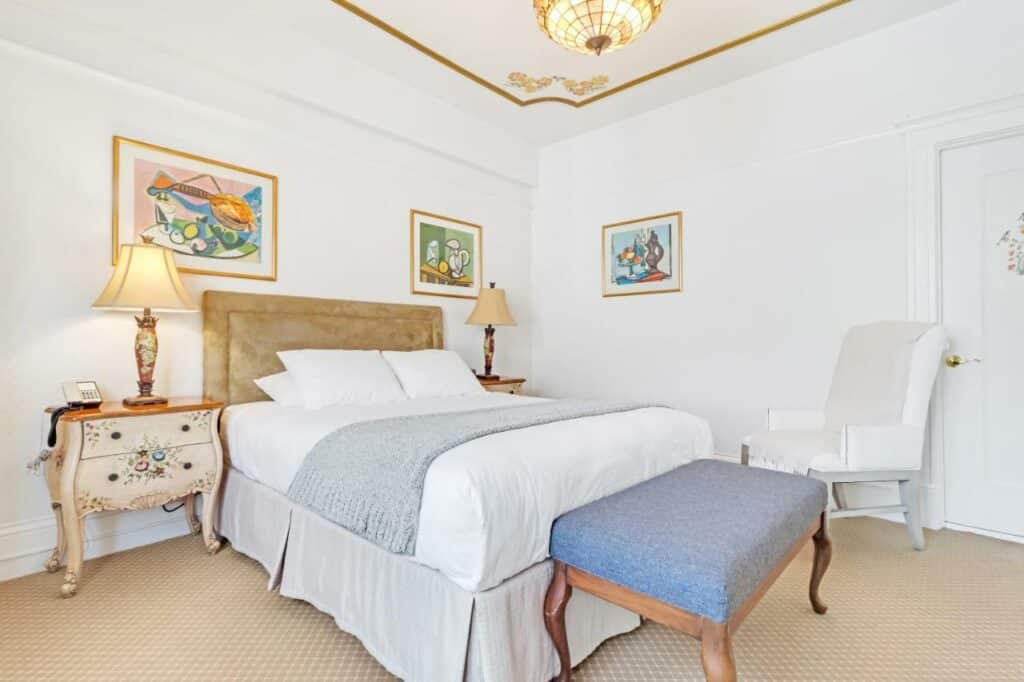 Quarto do Cornell Hotel de France com cama de casal do lado esquerdo da imagem com uma cômoda em cada lado da cama com luminária no pé da cama uma poltrona. Representa hotéis em San Francisco.