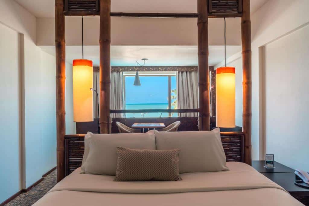 Quarto Eco Floor Luxo, com uma cama de casal com cabeceira feita em bambus, e atrás da cama tem uma mesa abaixo da janela que tem vista do mar