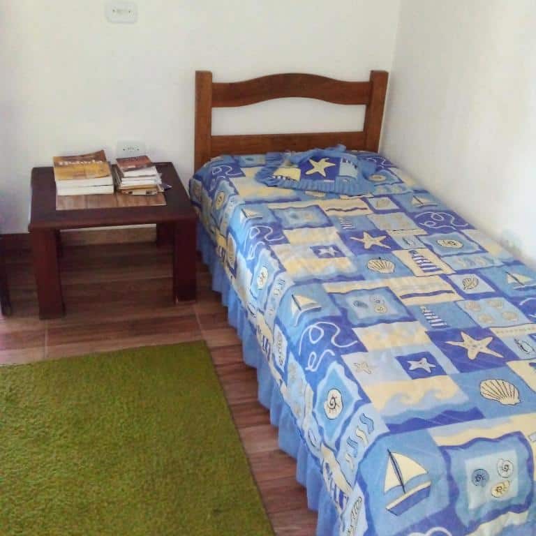 Quarto do Estalagem Sitio Colibri com uma cama de casal na parede direita da foto e do lado um tapete no chão e uma mesinha de madeira com livros.