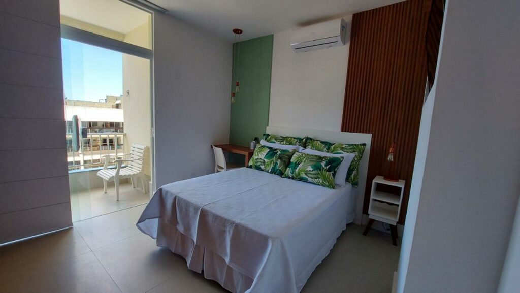 Quarto do Hospedagem de luxo no Barra Palace – Rio Paraíso com cama no centro do quarto, do lado direito da cama uma cômoda com luminária e do lado esquerdo uma mesa com cadeira. Representa airbnb na Barra da Tijuca.