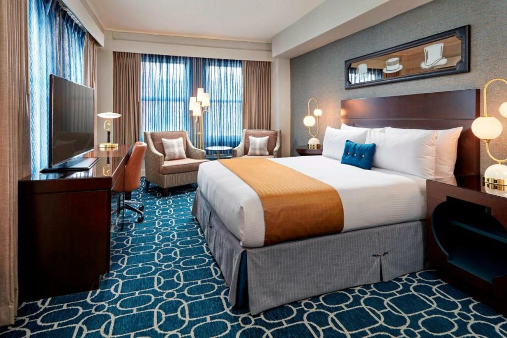 Quarto do Hotel Adagio, Autograph Collection com cama de casal do lado direito da imagem, com uma cômoda em cada lado da cama com luminária em frente a cama uma cômoda com TV e do lado esquerdo da cama duas poltronas. Representa hotéis em San Francisco.