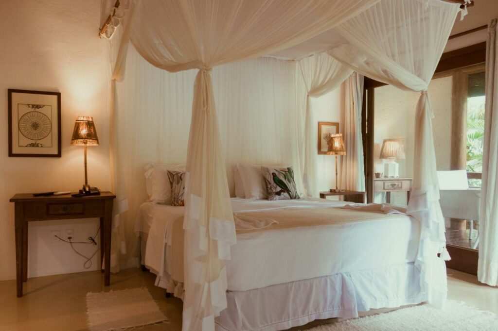 Quarto da Casa Hotel IFè Trancoso, próximo ao Quadrado com cama de casal no centro do ambiente e em cada lado da cama uma cômoda dom luminária.