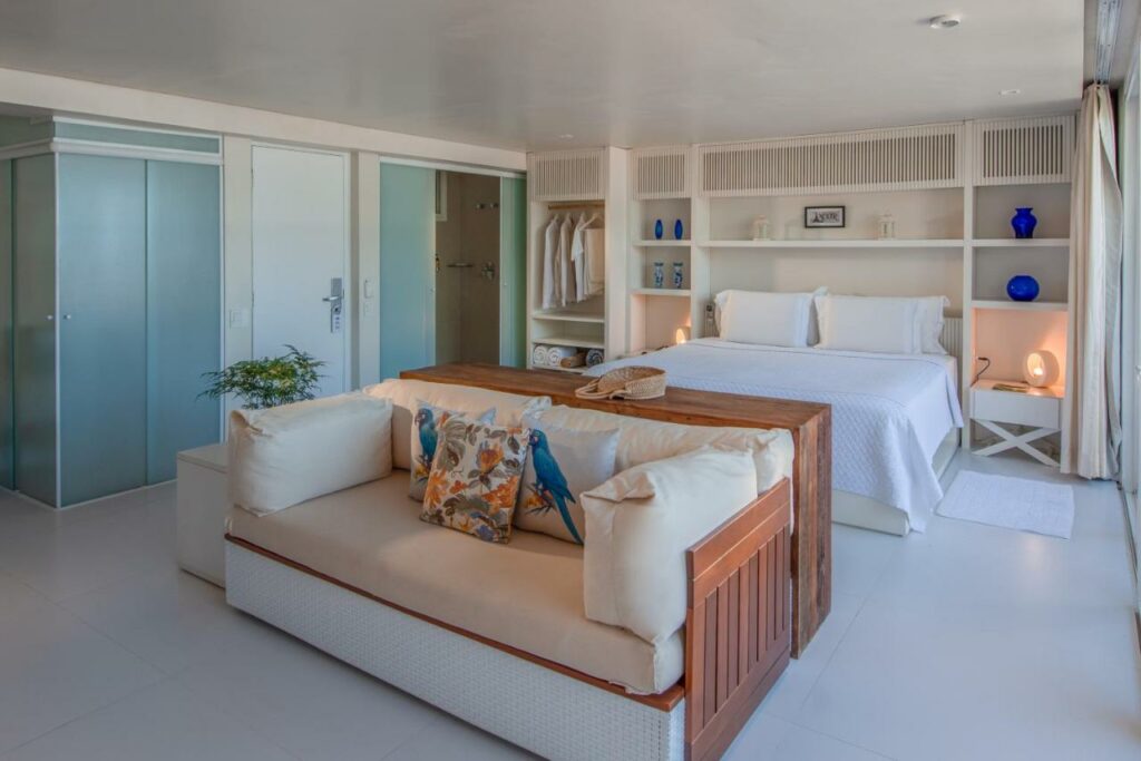 Quarto do Hotel Spa Nau Royal com sofá a frente do lado direito da imagem e ao fundo uma cama de casal com uma cômoda em cada lado da cama com luminária. Representa airbnb na praia da Juréia.