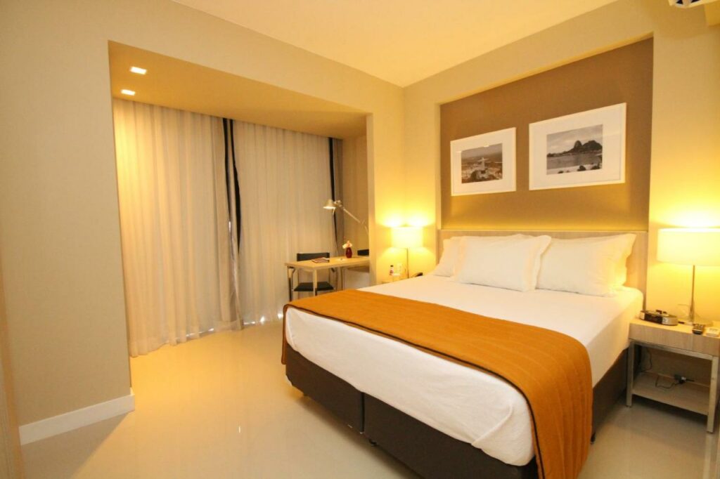 Quarto do Link Stay Flats com cama de casal do lado direito da cama com uma cômoda em cada lado da cama com luminária e do lado esquerdo da cama uma mesa de trabalho com cadeira. Representa airbnb na Barra da Tijuca.