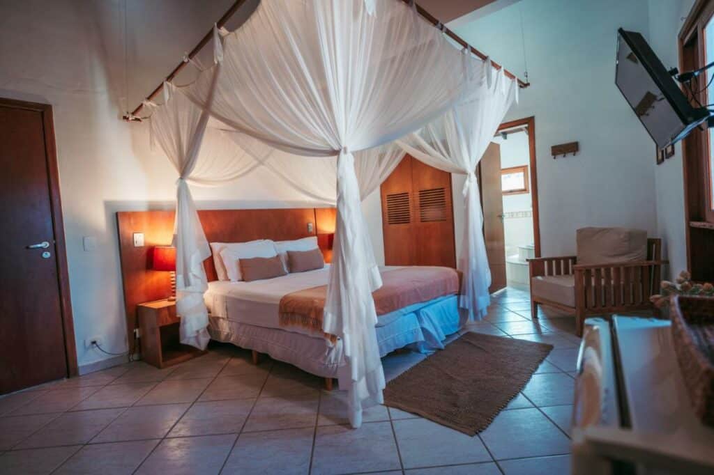 Quarto da Pousada Brigitte com uma cama de casal do lado esquerdo da imagem com uma cômoda em cada lado com luminária, do lado esquerdo da cama em frente uma poltrona. Representa airbnb na Barra do Sahy.