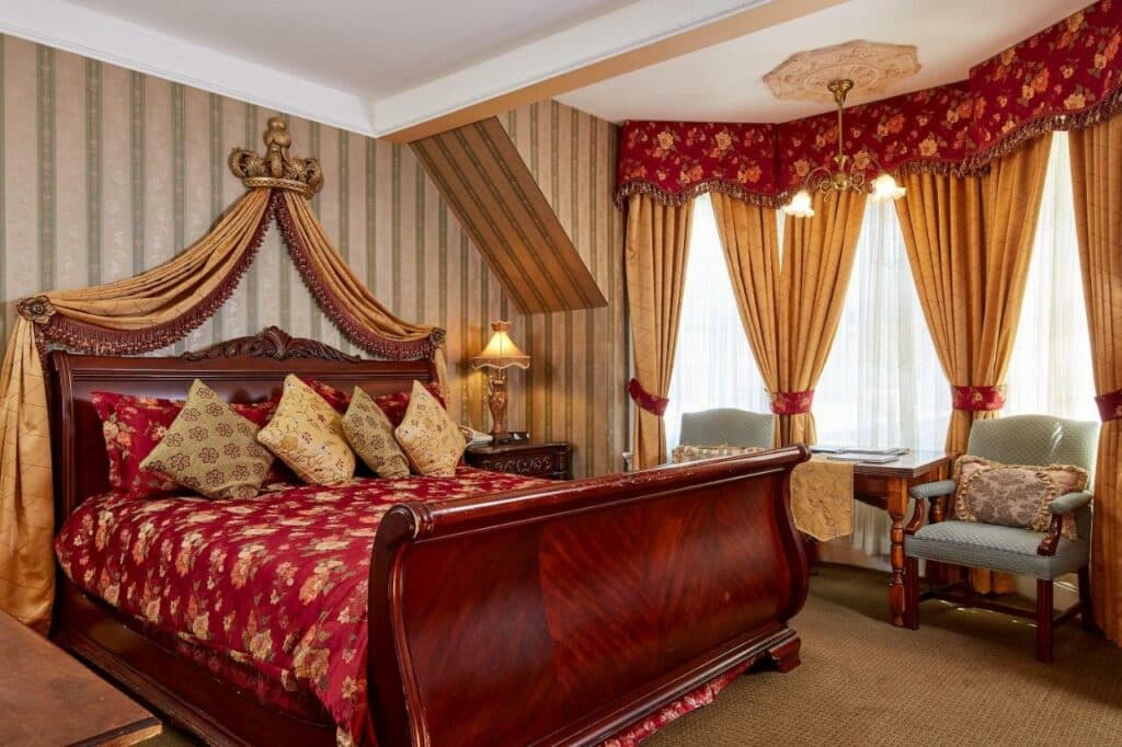 Quarto do Queen Anne com cama de casal do lado esquerdo da imagem, do lado esquerdo uma cômoda com luminária e do lado esquerdo da cama duas poltronas. Representa hotéis em San Francisco.