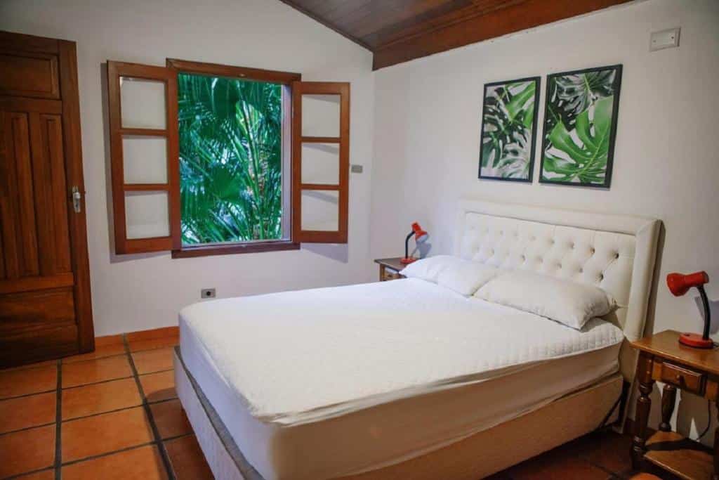 Quarto do Refúgio Toque Toque Chalés com cama de casal do lado direito da imagem com uma cômoda em cada lado da cama com luminária. Representa airbnb na praia de Calhetas.