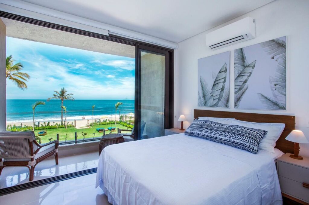 Quarto do Residencial Marina Del Sol com cama de casal do lado direito da imagem com uma cômoda em cada lado com luminária e do lado esquerdo da cama porta de vidro com acesso a varanda com vista para o mar. Representa airbnb na praia de Calhetas.