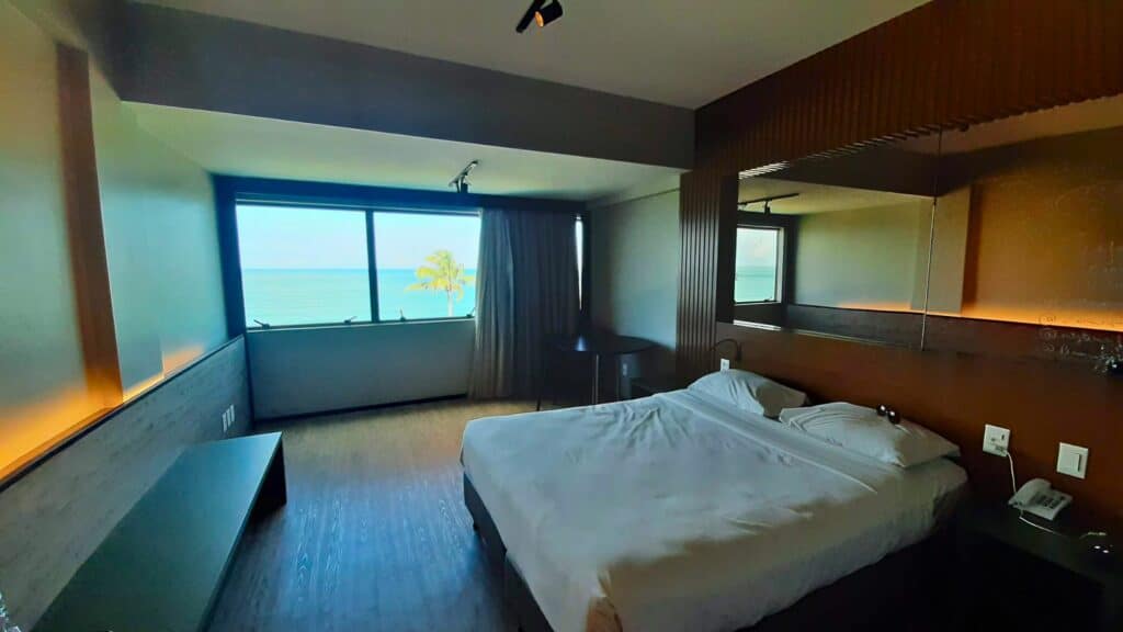 Quarto Premium Plus Luxo do Ritz Lagoa da Anta, com uma cama de casal, um espelho retangular acima da cama, uma mesa preta com duas cadeiras (à esquerda da cama) e uma janela ampla com vista do mar e de um coqueiro.