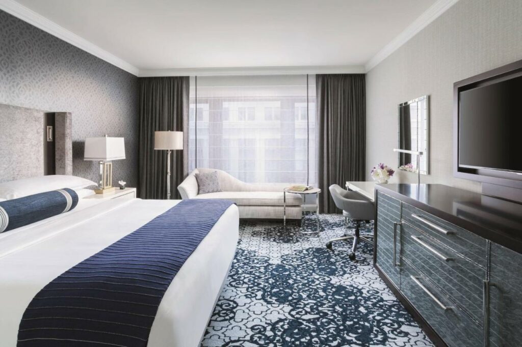 Quarto do The Ritz-Carlton, San Francisco com cama de casal do lado esquerdo da imagem e em frente a cama uma cômoda com TV e do lado esquerdo da cama uma sofá. Representa hotéis em San Francisco.