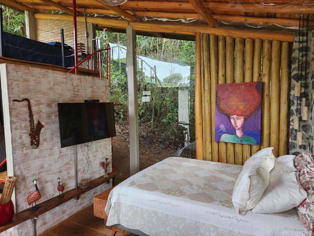 Quarto de 36m² do Tiny Houses O Olival - aluguel temporada com uma cama de casal e em frente uma parede com vários objetos e uma tv. Do lado da cama tem um parede de madeira com um quadro e do lado uma parte de vidro.