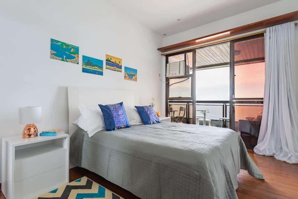 Quarto do Vista DE CINEMA do 19 andar da Praia BARRA da TIJUCA – Portaria 24h, Estacionamento, Wi-Fi 35mbps, Ar Condicionado – BANHEIRO RECEM REFORMADO com cama de casal do lado esquerdo da imagem com uma cômoda em casa lado da cama e do lado esquerdo da cama uma porta de vidro com acesso a varanda.