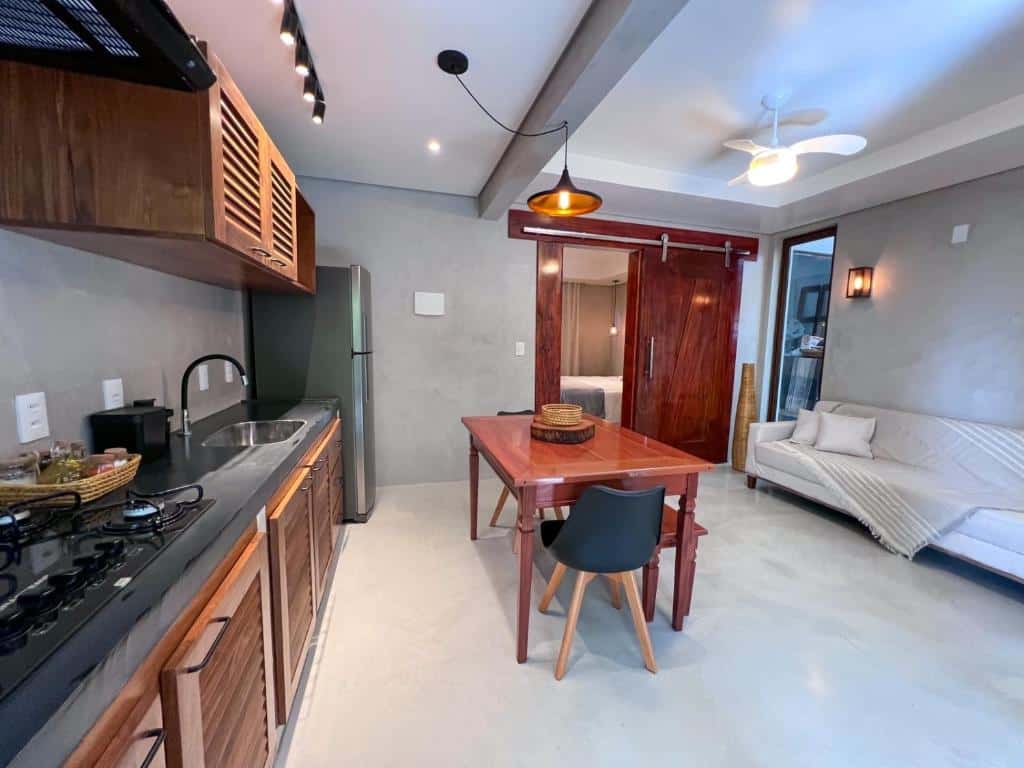 Cozinha do Refugio Do Morro. Do lado esquerdo o fogão, a pia, os armários e a geladeira. No meio uma mesa de madeira com cadeiras. Do lado direito um sofá, no fundo a porta do quarto.