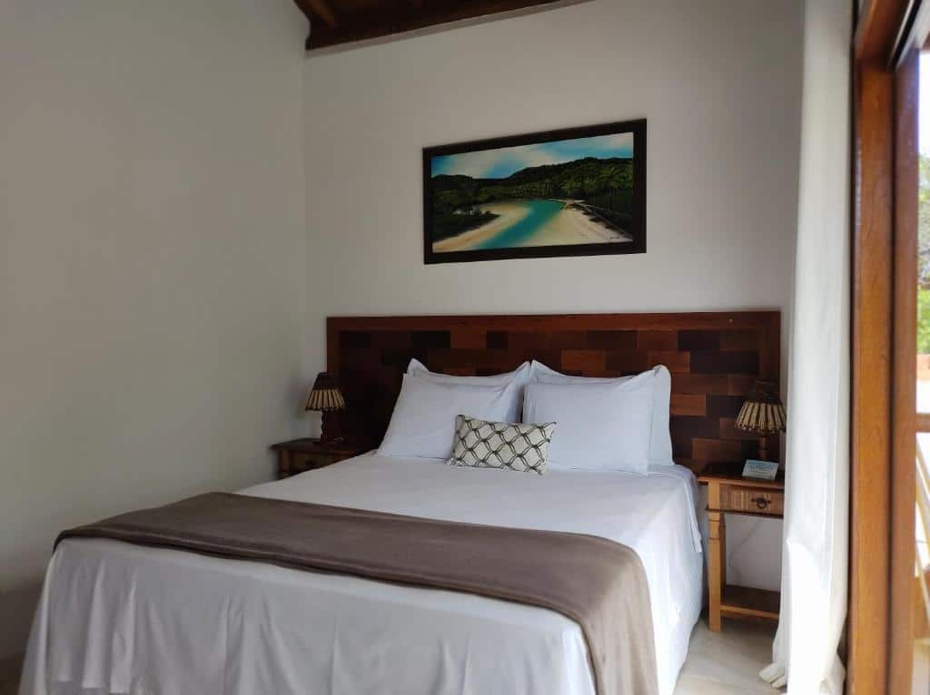 Quarto do Conchas do Mar Residence. A cama box de casal com mesas de cabeceira está no meio do ambiente. Atrás dela há um quadro na parede. Á direita há uma varanda. Representa o post sobre airbnb em Itacaré.