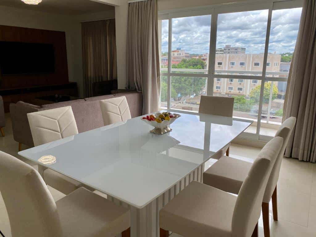 Área de refeições no RESIDENCIAL SHAFA. A mesa contém seis cadeiras e está de frente para uma varanda com vista. No fundo à esquerda vemos parte da sala de estar.