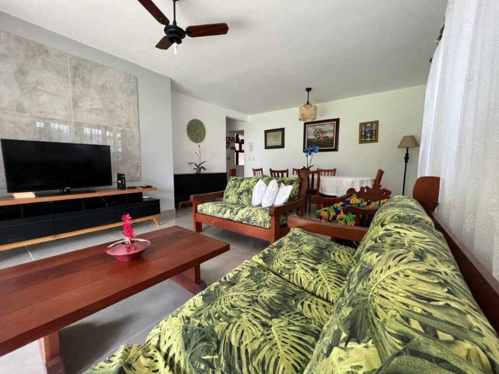 Sala de estar da Casa Vivá Guaeca com sofá do lado direito da imagem com uma mesa de centro e a frente uma cômoda com TV.