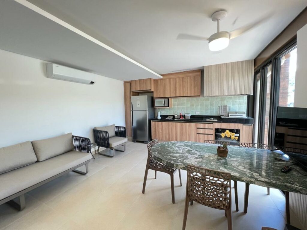 Cozinha do Flores de Juquehy com mesa de mármore do lado direito da imagem com cadeiras, do lado esquerdo um sofá e ao fundo do ambiente armário, com geladeira, micro-ondas, pia e fogão.