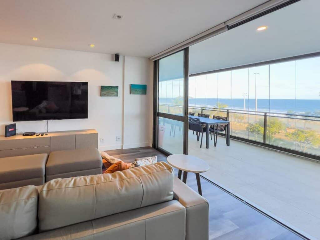 Sala de estar do Luxo e Vista Mar Maravilhosa na Barra da Tijuca B11-0013 com sofá e TV do lado esquerdo da imagem do lado direito uma porta ampla aberta com acesso a varanda.