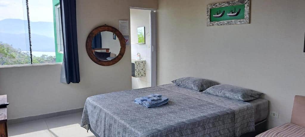 Quarto da Suíte em casa particular. Uma cama de casal do lado direito, no fundo um espelho e a porta do banheiro. Do lado esquerdo uma janela de vidro com vista para o mar. Foto para ilustrar post sobre airbnb na Praia da Almada em Ubatuba.