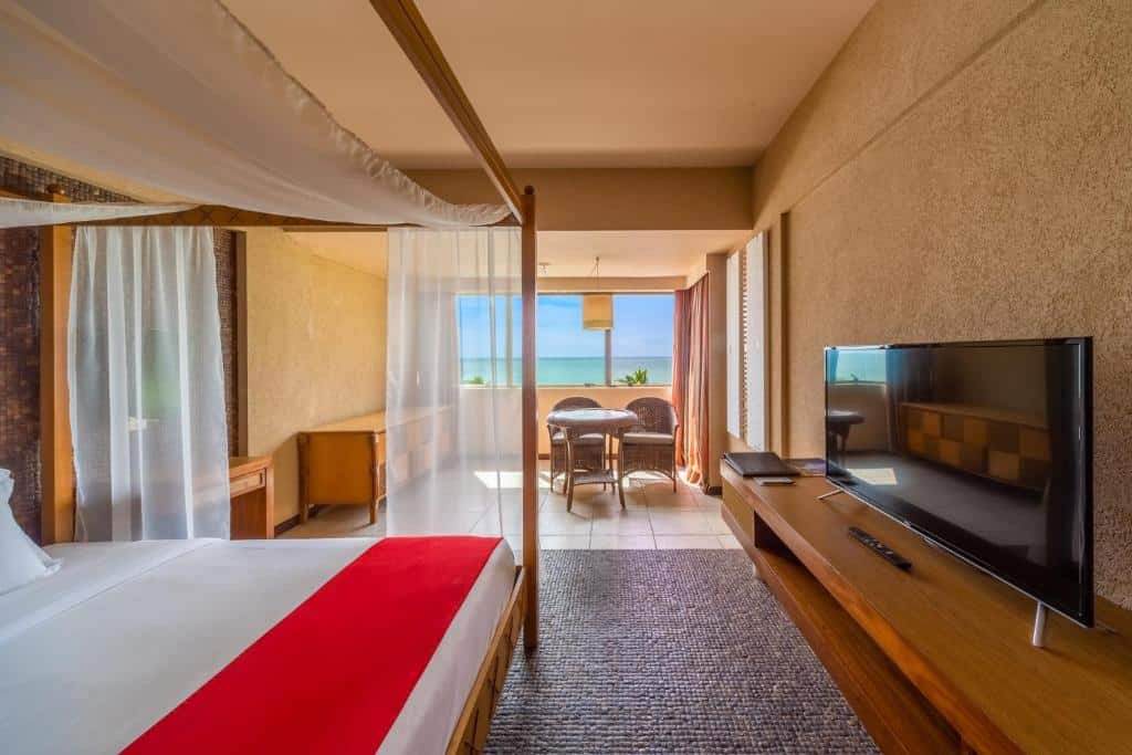 Suíte superior localizada no andar Bali do Ritz Lagoa da Anta. Há um cama de casal com dossel, móvel à frente com TV em cima, e uma mesa com duas cadeiras e janela ampla com vista do mar