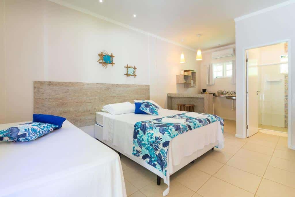 Quarto do Suites Prime. Uma cama de solteiro e uma de casal do lado esquerdo, no fundo um balcção com bancos e uma pia com utensílios de cozinha. Do lado direito a porta do banheiro.