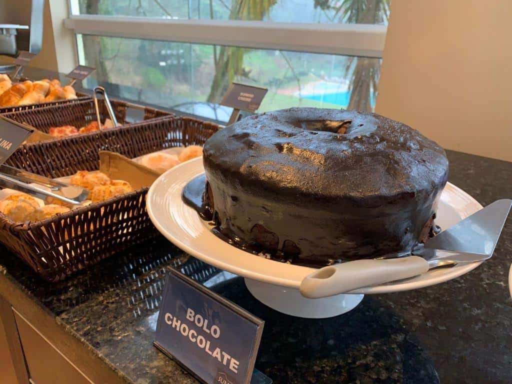 Buffet de café da manhã no Summit Inn Hotel. Vemos um bolo de chocolate e cestas com pães e massas.
