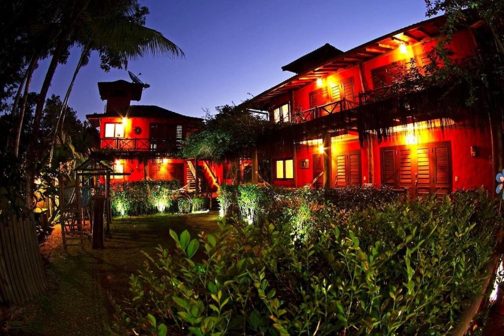 Área externa do Thai Suítes Itamambuca. Do lado esquerdo um parquinho de madeira infantil. do lado direito os quartos com rede na frente. Na frente um jardim a noite todo iluminado.