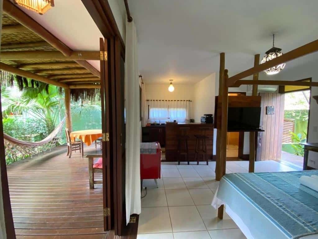 Quarto do Thai Suítes Itamambuca. Do lado direito uma cama de casal, de frente uma televisão e um frigobar. No fundo a porta do banheiro e uma mini cozinha. Do lado esquerdo a varanda, com uma rede, uma mesa e um banco.