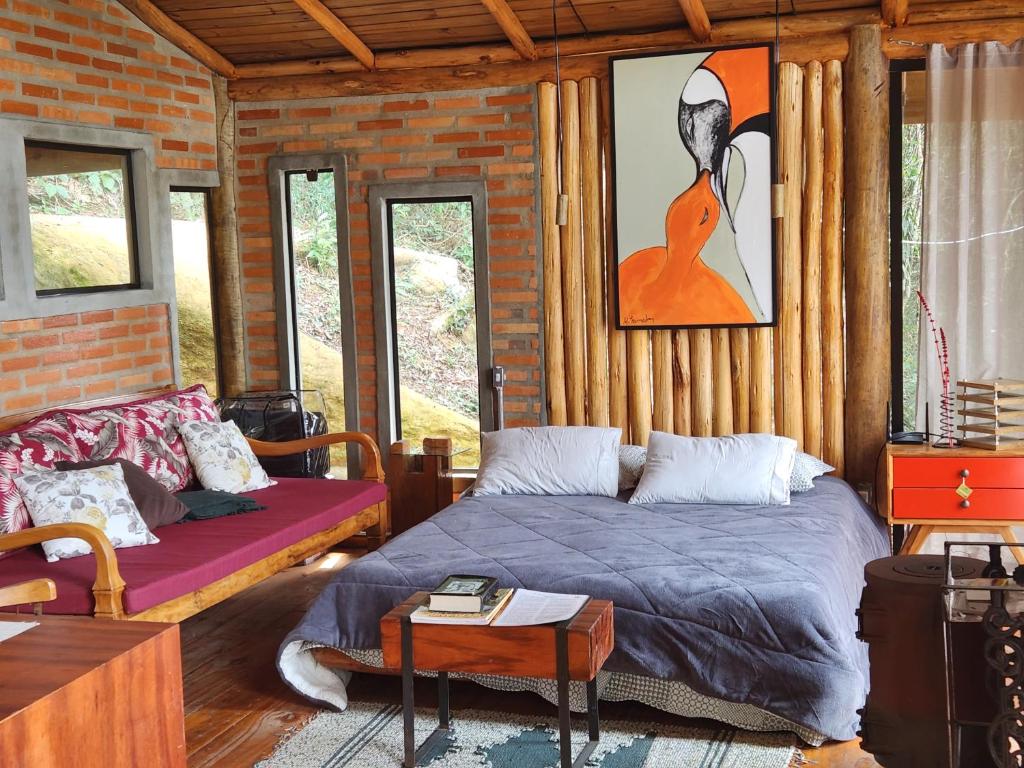 Quarto com 34m² do Tiny Houses O Olival - aluguel temporada com uma cama de casal atrás uma parede de madeira com um quadro. Do lado esquerdo da cama tem um sofá com almofadas e as paredes são de tijolos a vista com janelas e portas de vidro. Já do lado direito tem uma mesinha do lado da cama e outros objetos. Imagem ilustrando post Airbnb em Cunha.