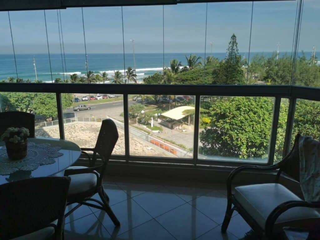 Varanda do Apartamento frente mar, Posto 5 – Barra da Tijuca com mesa e cadeiras do lado esquerdo da imagem e do lado direito uma cadeira com vista para o mar.