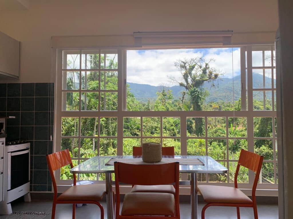 Cozinha da Vila Itamambuca Apto Passarinhos. Uma mesa na frente, atrás uma parede de vidro com janela e vista para as montanhas, do lado esquerdo a pia com fogão.